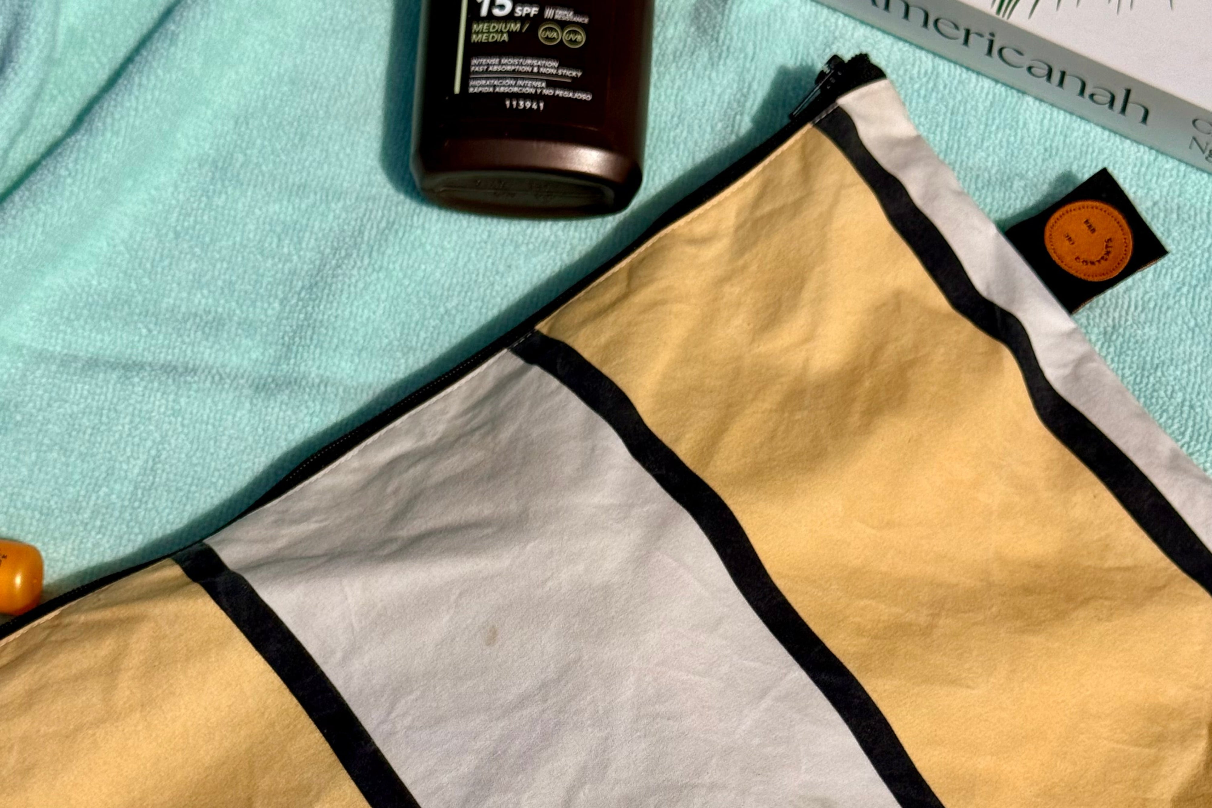 A striped beach towel or pouch in yellow, white, and black, laid out on a light blue towel with a brown bottle and a book nearby.