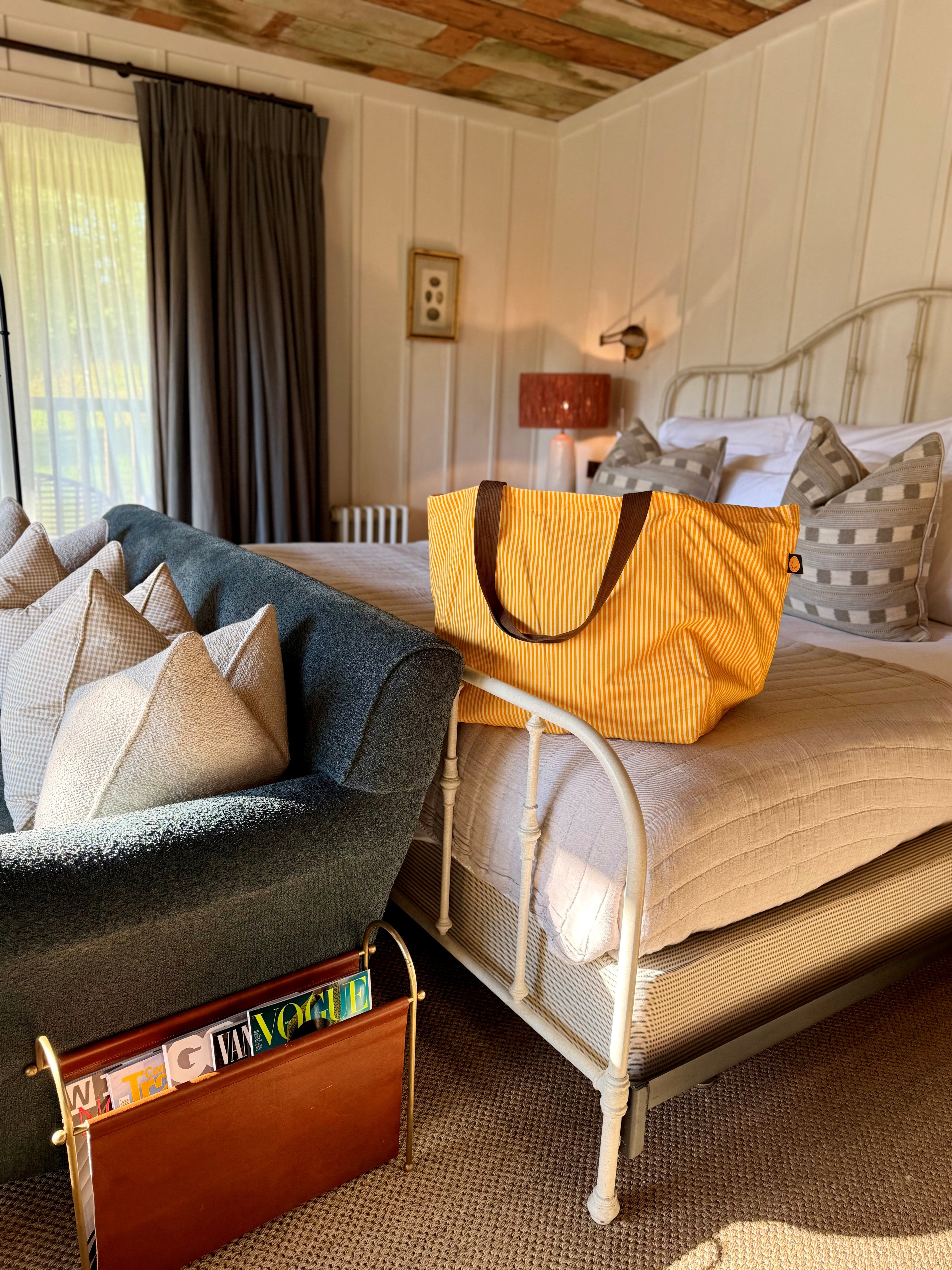 Yellow bag on a bed in a bedroom with a magazine rack and lamp.